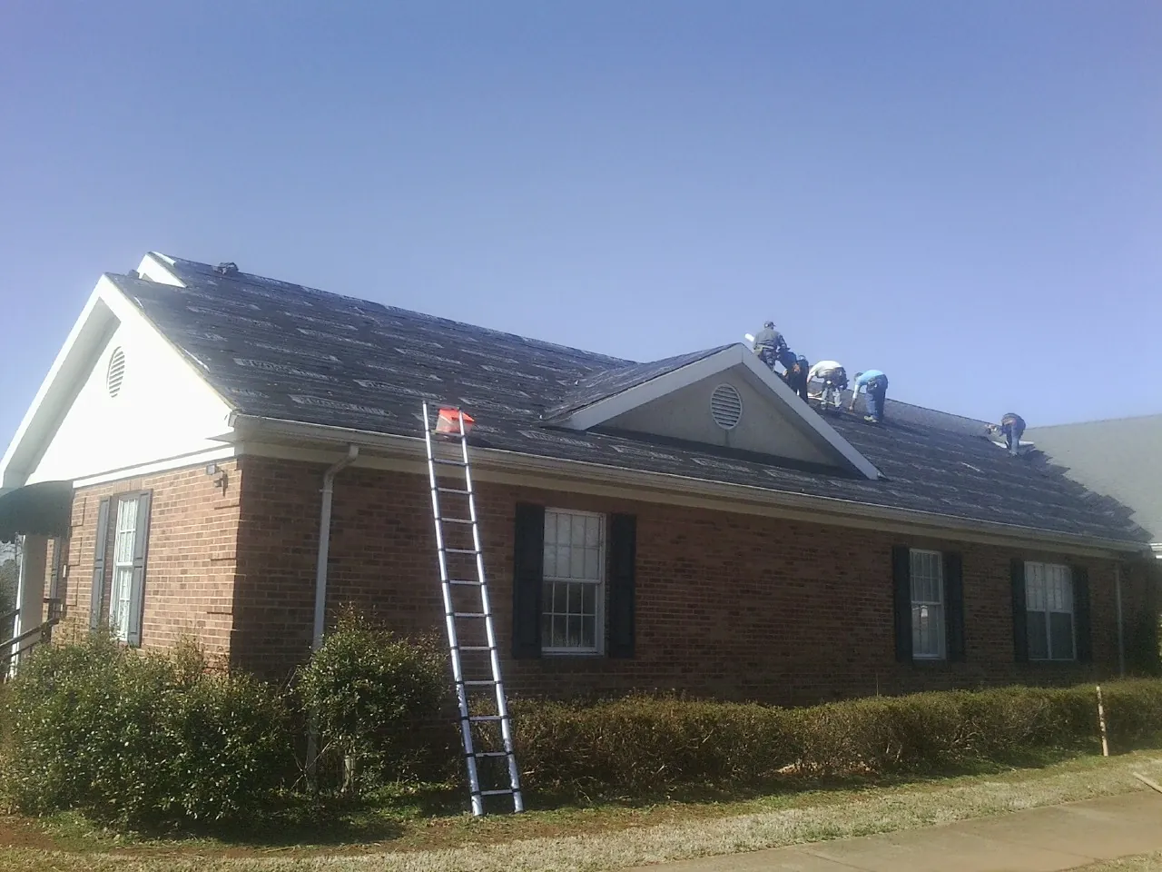 Crew tearing shingles off a brick ranch roof with a ladder set — Emergency Roof Repair work by our crew in Dracut
