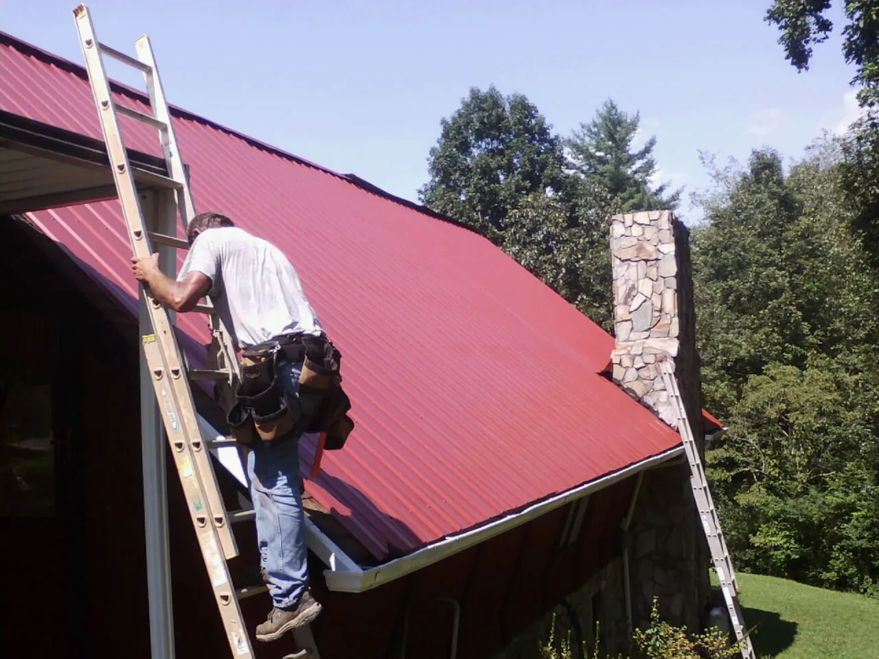 Roofer on a ladder beside a red metal roof and stone chimney — Soffit & Fascia Repair by Roofing Brothers in Dracut