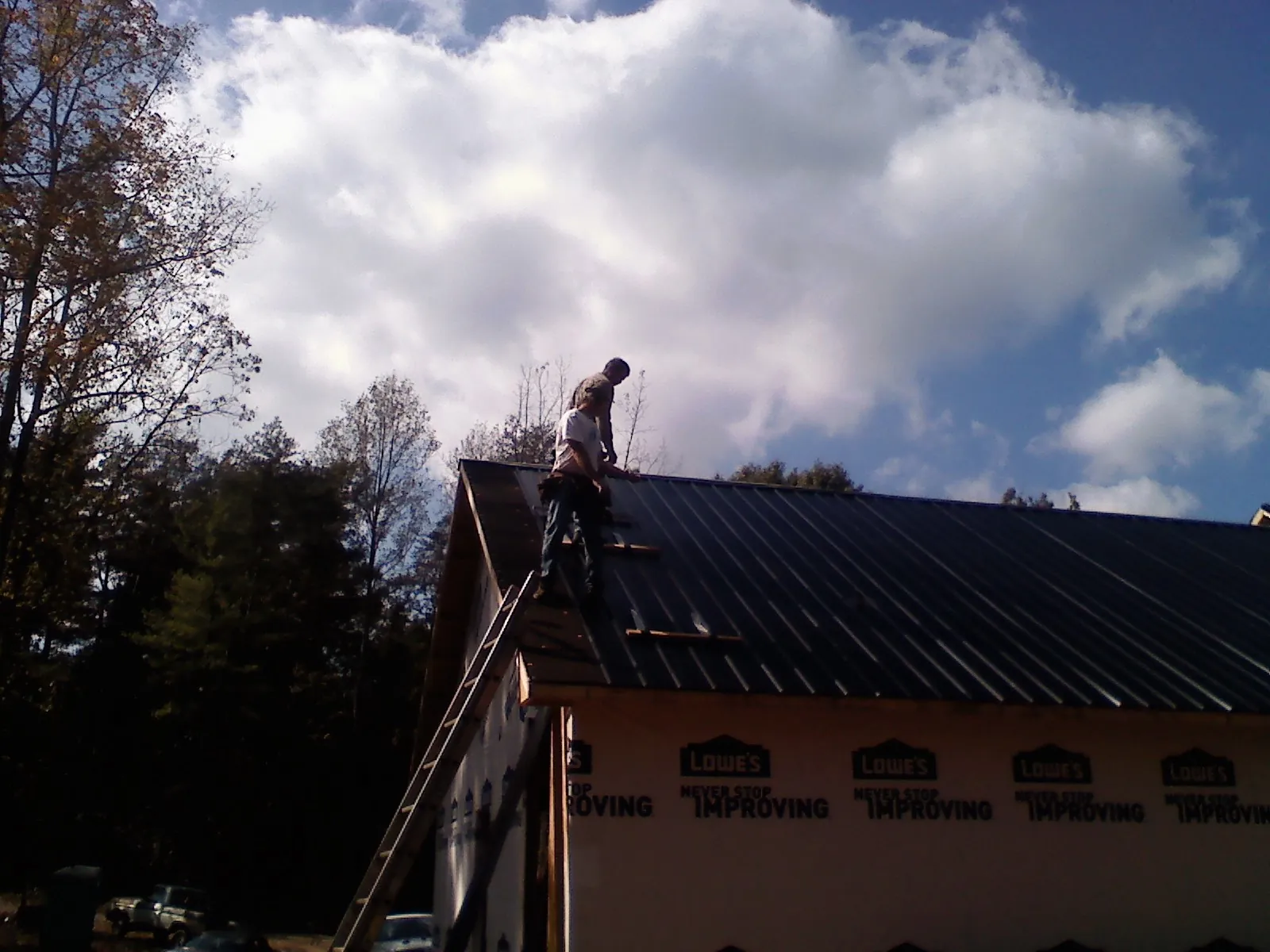 Roofer fastening dark metal panels high against a cloudy sky, from a Roof Inspection job in Dracut